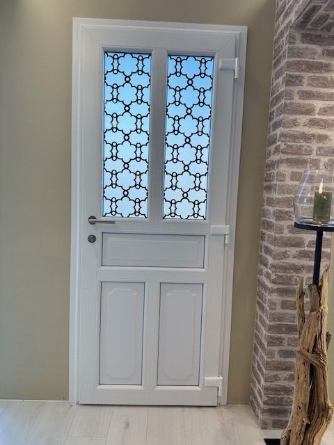 Porte d'entrée blanche avec panneaux de verre à motifs, adossée à un mur de briques dans un hall d'entrée beige.