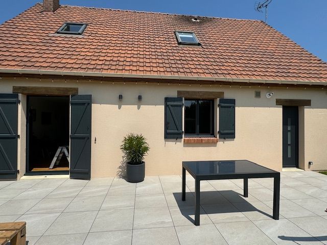 Extérieur d'une maison beige au toit de tuiles rouges, volets ouverts et table de patio sur une terrasse ensoleillée