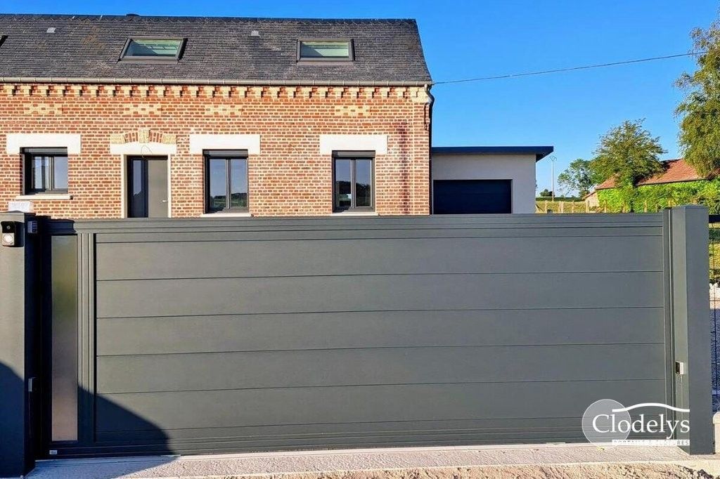 Maison moderne en briques derrière un haut portail coulissant gris foncé, sous un ciel d'un bleu limpide.