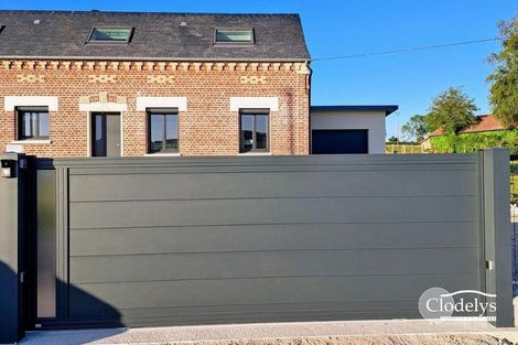 Maison en briques derrière un haut portail coulissant gris, sous un ciel bleu.