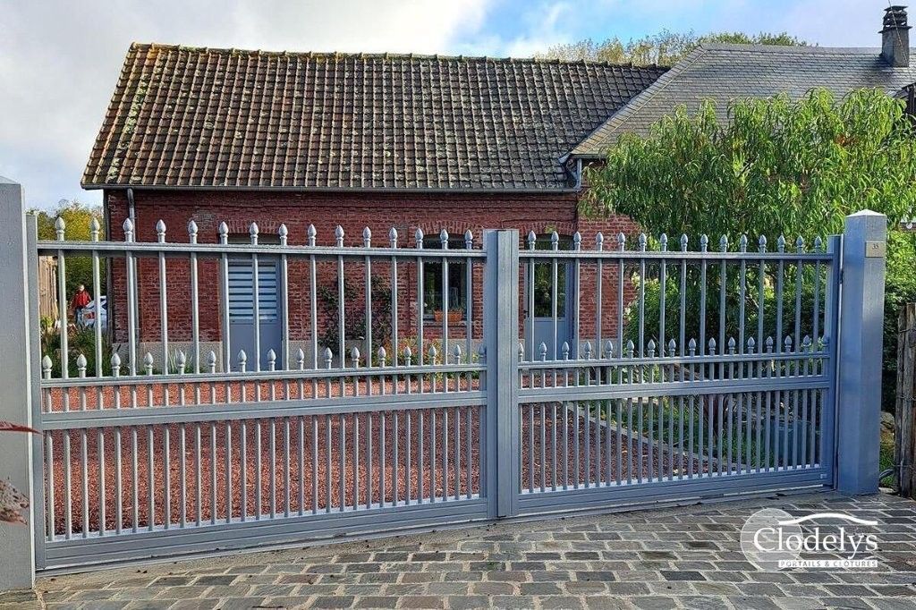 Maison derrière une clôture métallique grise et une rue pavée, avec un mur en briques rouges et un toit de tuiles.
