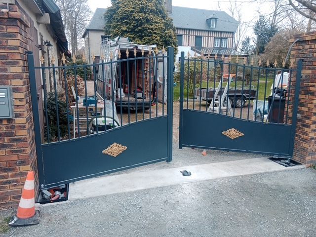 Portail d'allée en fer forgé noir en cours d'installation à l'entrée d'une maison, avec des cônes de signalisation à proximité