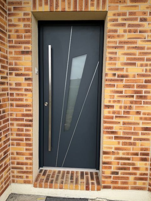 Porte d'entrée moderne grise avec un panneau de verre étroit encastré dans un mur de briques.