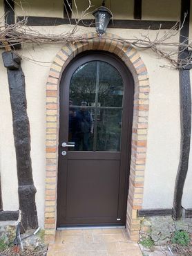 Porte cintrée brune avec encadrement en briques sous une petite lampe extérieure, encadrée par des poutres en bois recouvertes de vigne.