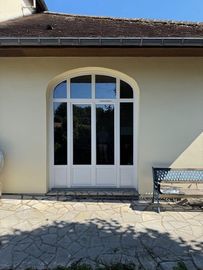 Double porte-fenêtre cintrée blanche sur une maison beige, avec des dalles de terrasse et un banc à proximité.