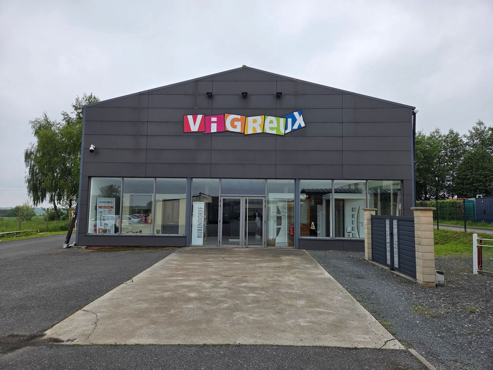 Vue de face d'un bâtiment gris avec une enseigne colorée au-dessus de portes d'entrée vitrées et une allée en béton.