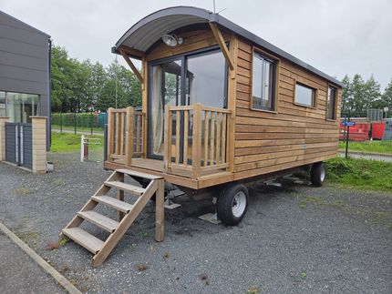 Petite cabane en bois sur roues avec porche et marches, garée à l'extérieur sur du gravier.