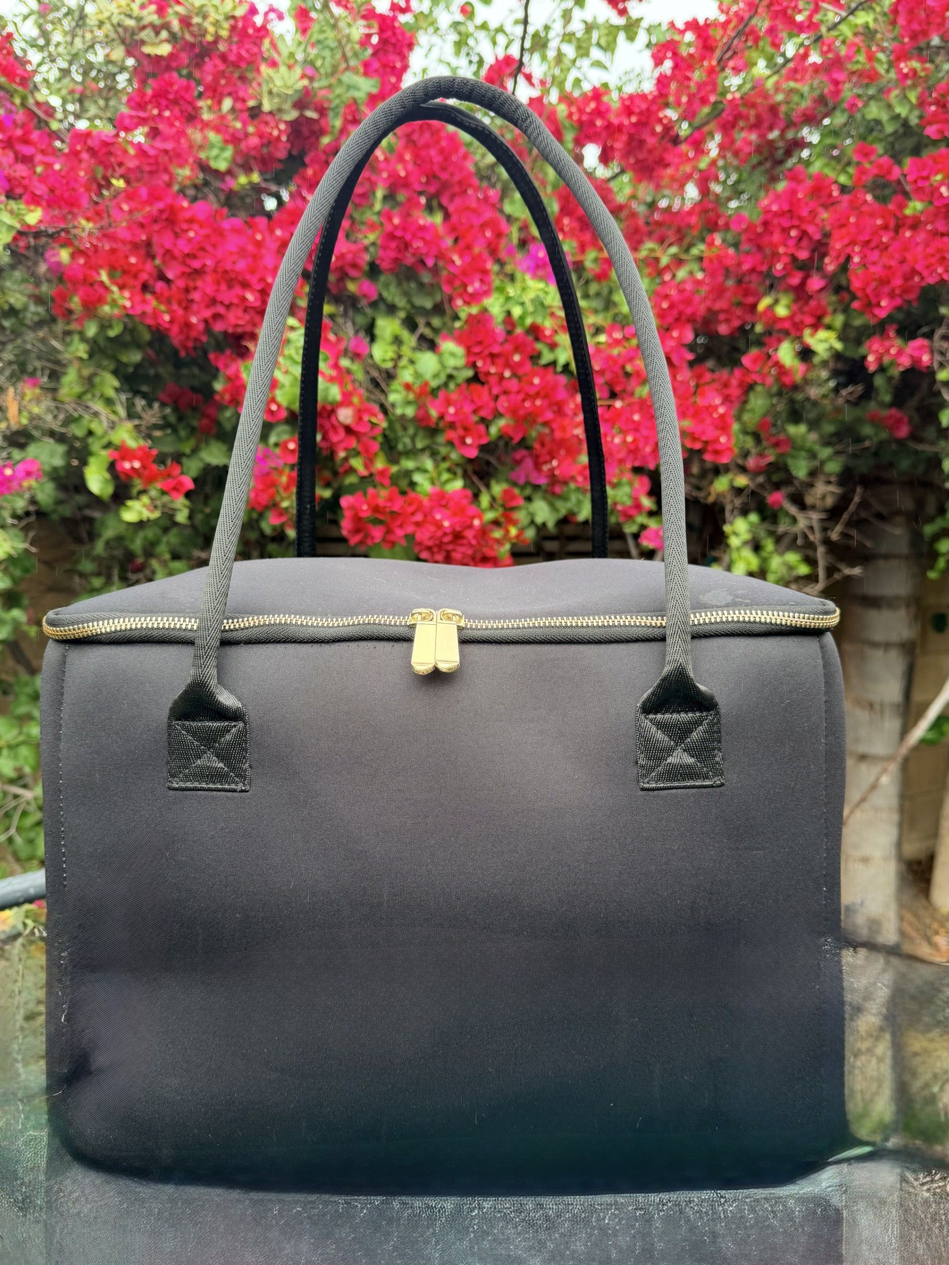 The Birdie black insulated tote bag by The Stever Co. with gold zipper accents displayed outdoors in front of bright red flowers.