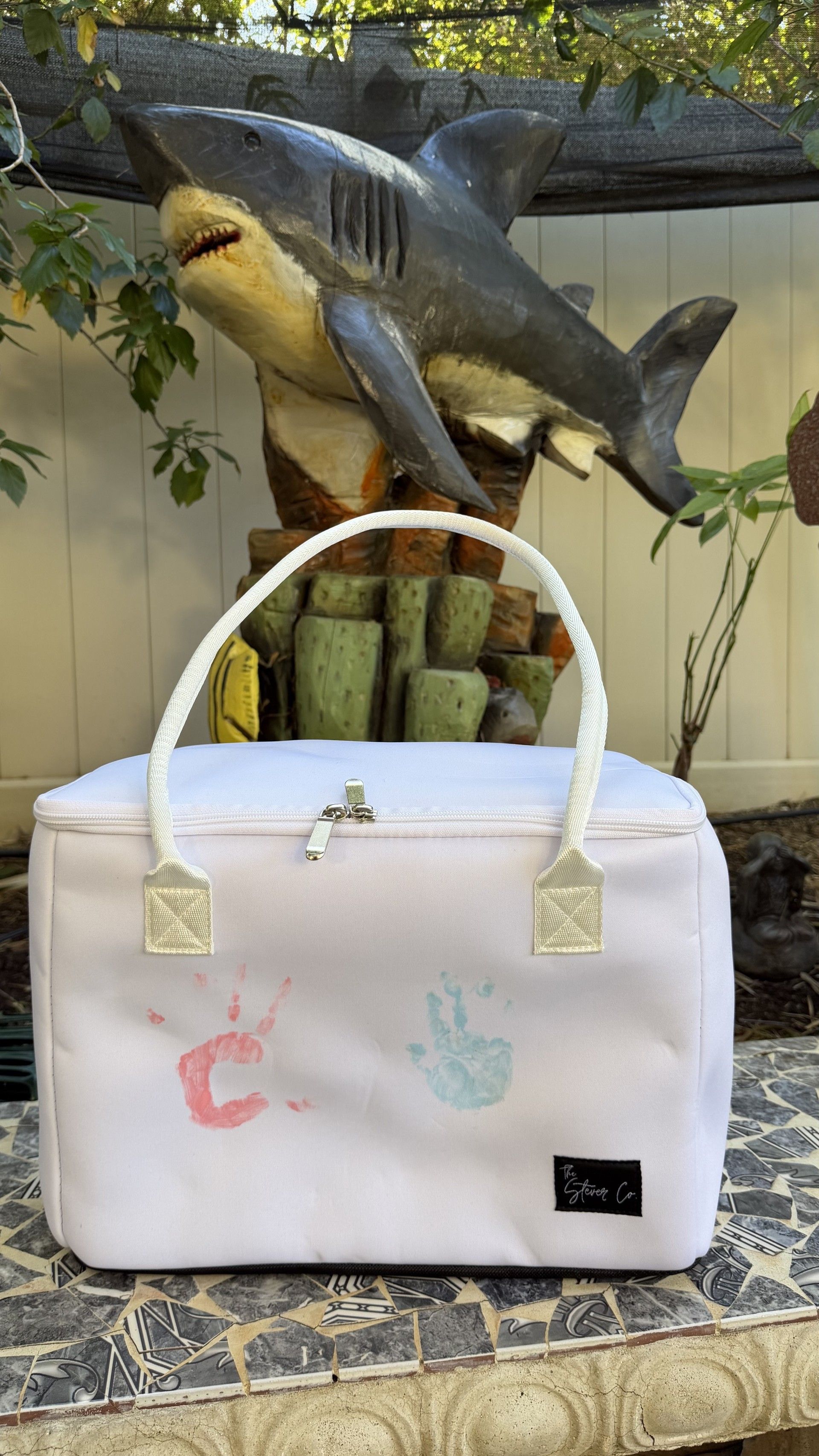 Mom’s Favorite white insulated tote bag by The Stever Co. decorated with children’s handprints and displayed outdoors with a shark sculpture in the background.