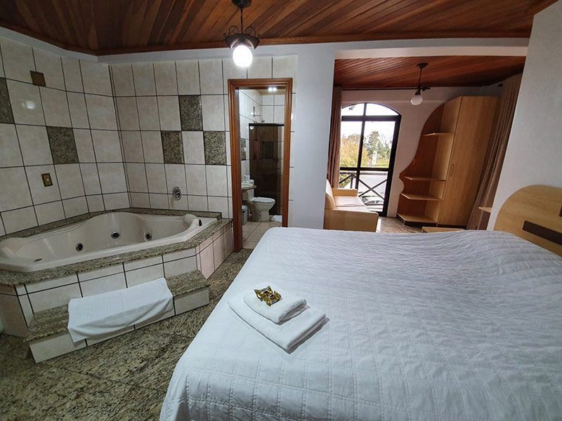 Quarto de hotel com uma cama grande, uma banheira de hidromassagem no canto e uma porta que dá acesso a um pequeno banheiro e varanda.