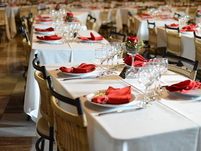Fileiras de mesas de banquete postas com toalhas de mesa brancas, taças de vinho e guardanapos vermelhos dobrados em um salão de jantar formal.