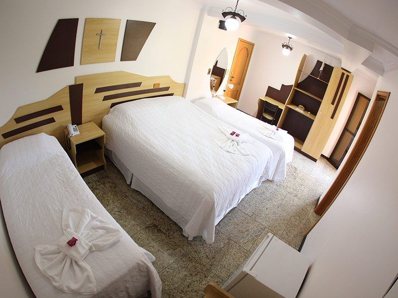Um quarto de hotel com uma cama de solteiro e uma cama de casal, roupa de cama clara, cabeceiras de madeira e um armário alto.
