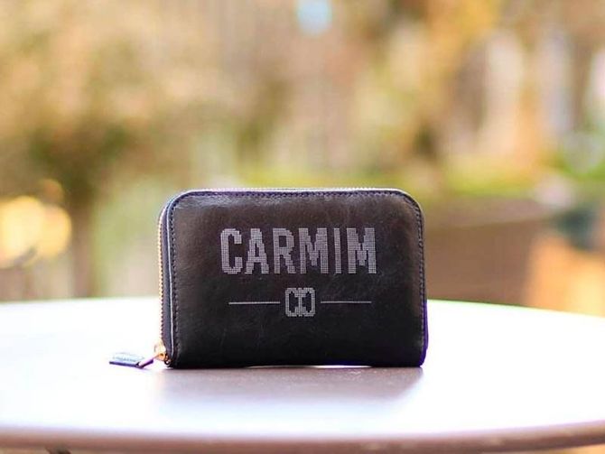 Uma carteira retangular cinza-escura com zíper, com a inscrição "CARMIM" e um pequeno emblema impressos, sobre uma mesa redonda ao ar livre.