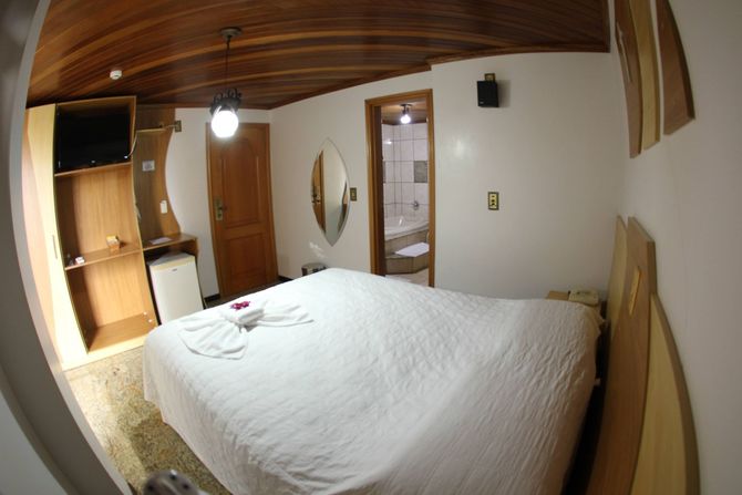 Um quarto de hotel com cama de lençóis brancos, teto de madeira, guarda-roupa, frigobar e porta que dá acesso ao banheiro.