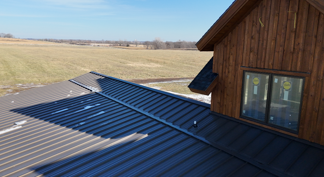 Dark metal roof with a wood-sided building, field in the background under a blue sky.