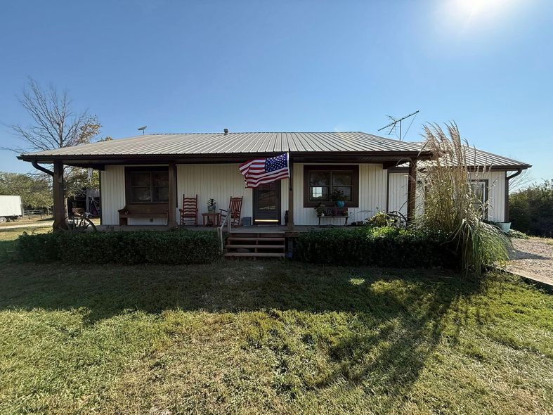 White house with covered porch, American flag, green yard, blue sky, sunny day.