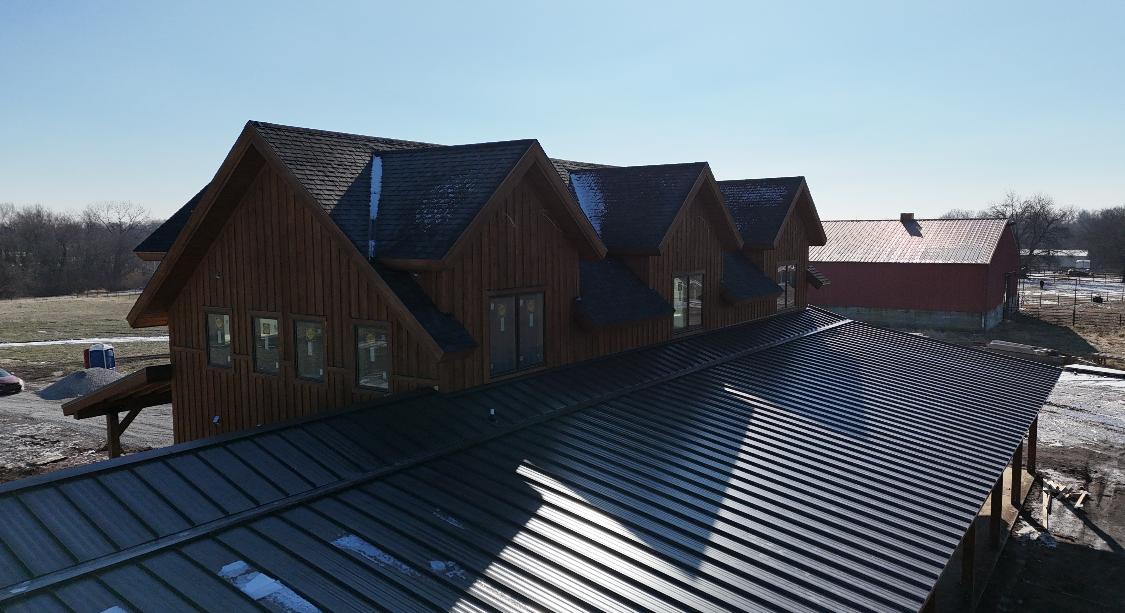 Brown wooden building with multiple gabled roofs, a long curved metal roof, and a red barn in the background.