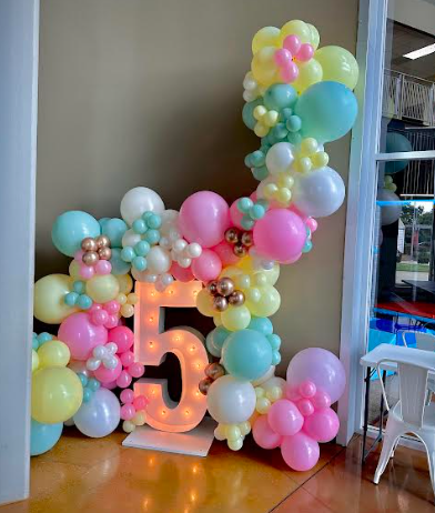 A colorful balloon arch with a lit-up number 5. Pastel pink, blue, and yellow balloons decorate a room.