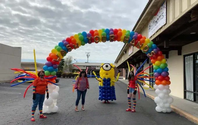 People in costumes pose under a rainbow balloon arch. A large minion stands in the middle.
