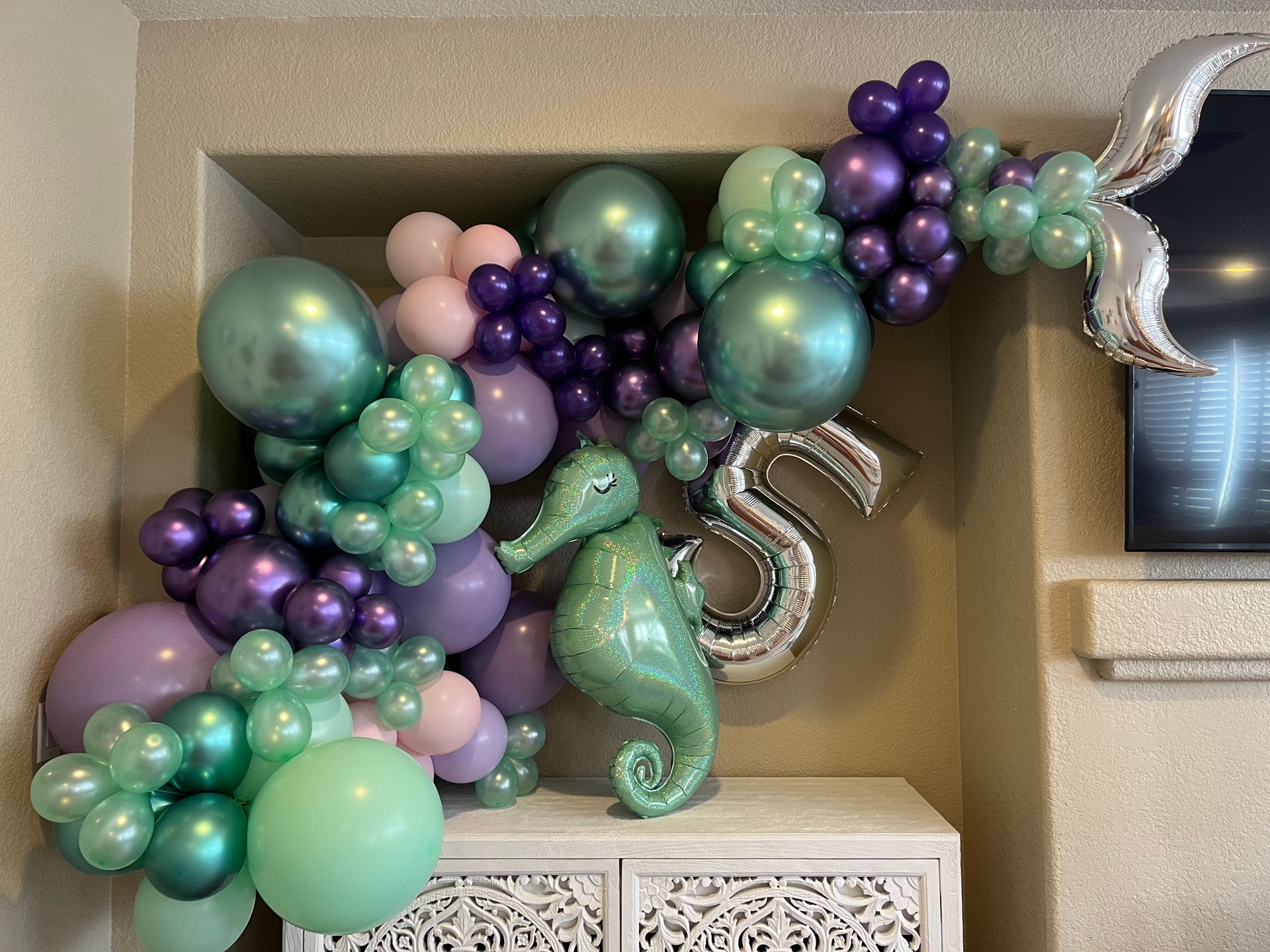 Mermaid-themed birthday decoration with balloons in shades of purple, green, and silver, plus a seahorse and number 5.