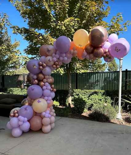 Balloon arch in shades of purple, peach, and rose gold; set outdoors with a tree and greenery.