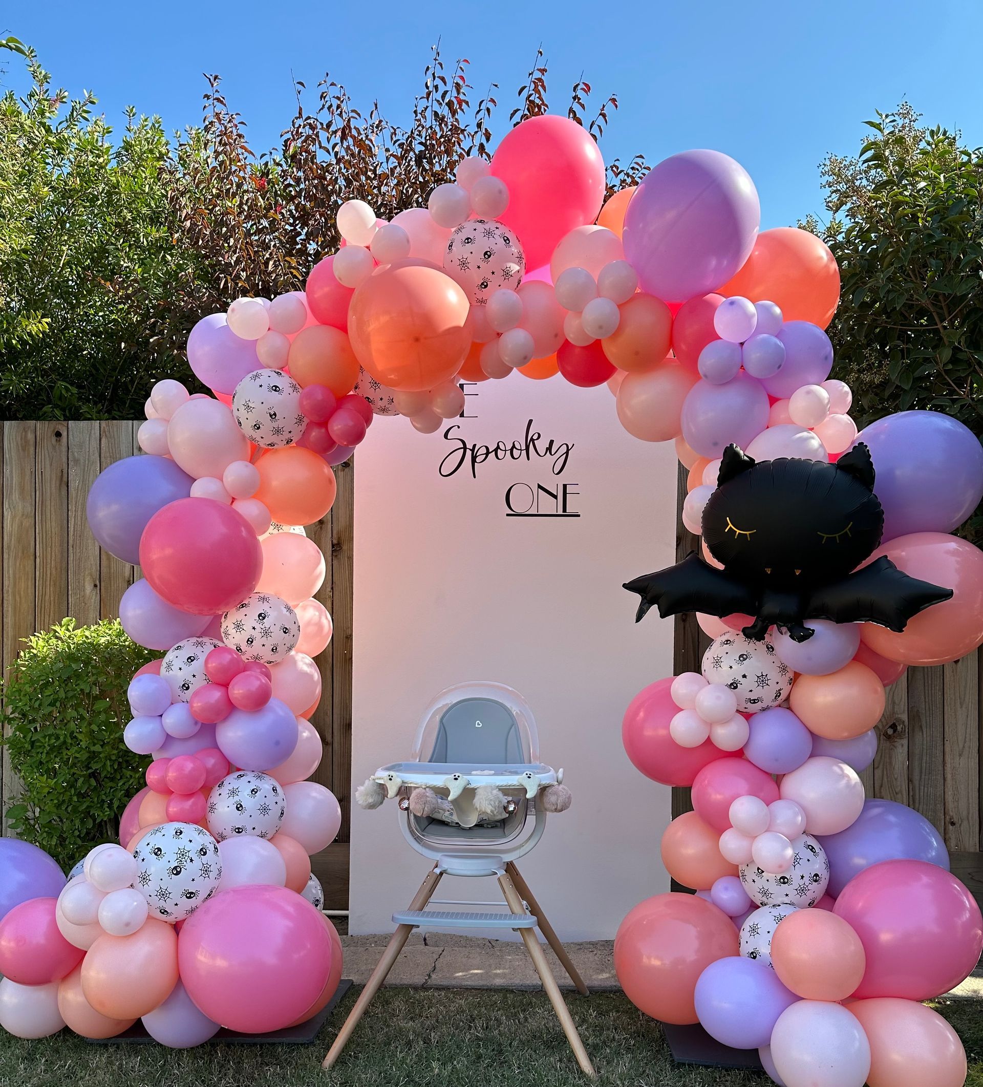 A festive outdoor birthday setup with balloon arch, highchair, and bat decoration.
