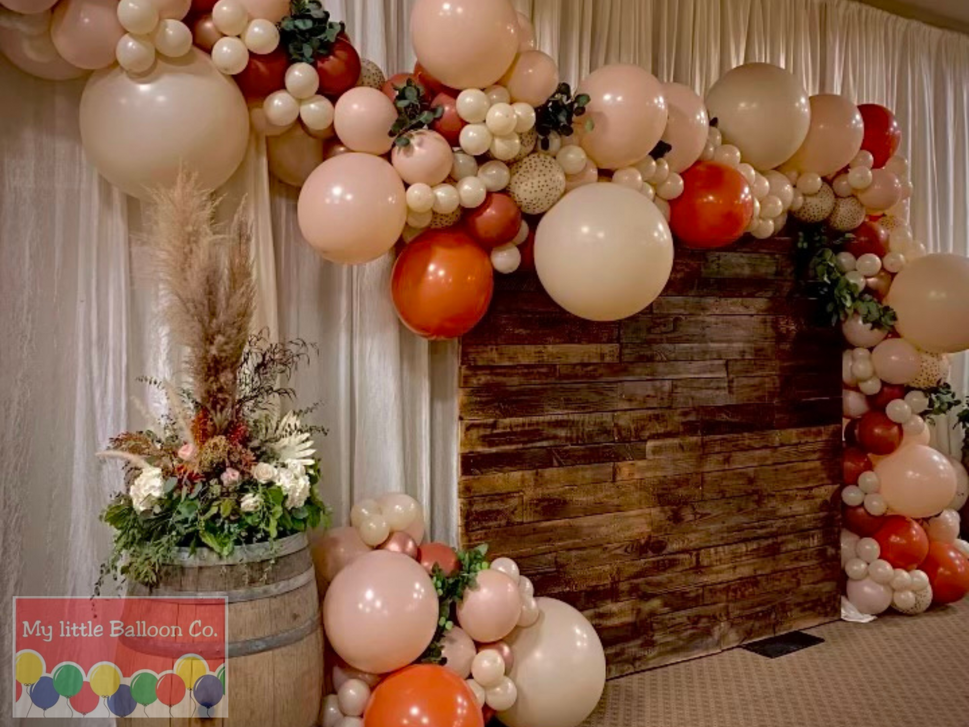 Balloon arch with wood backdrop, floral arrangement in a barrel. Neutral and orange balloons.