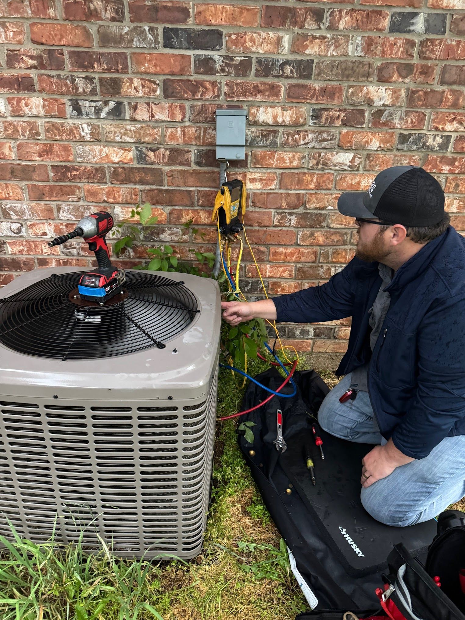 Technician kneels beside an outdoor HVAC unit, checking gauges and hoses against a brick wall.