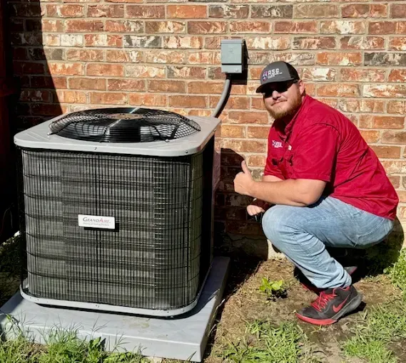 Technician kneeling beside outdoor air conditioner unit against a brick wall.