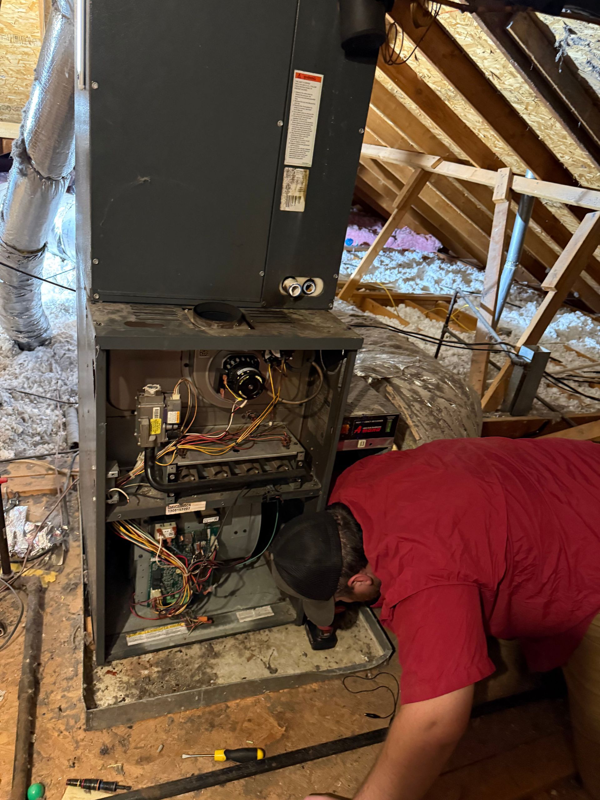 Technician kneeling by an open furnace in an attic, inspecting exposed wiring and components.