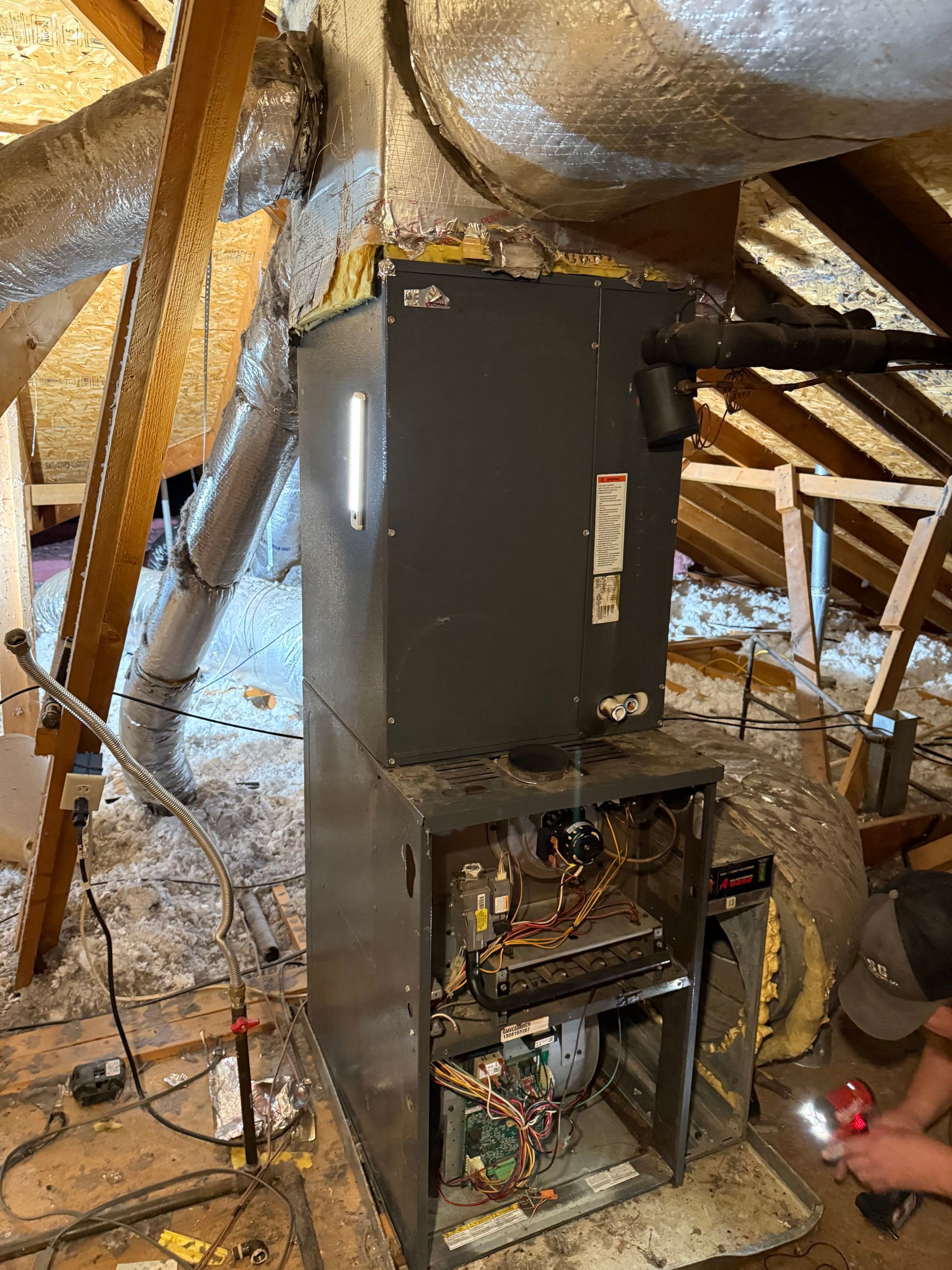 Black HVAC furnace in an unfinished attic with exposed insulation and open access panel