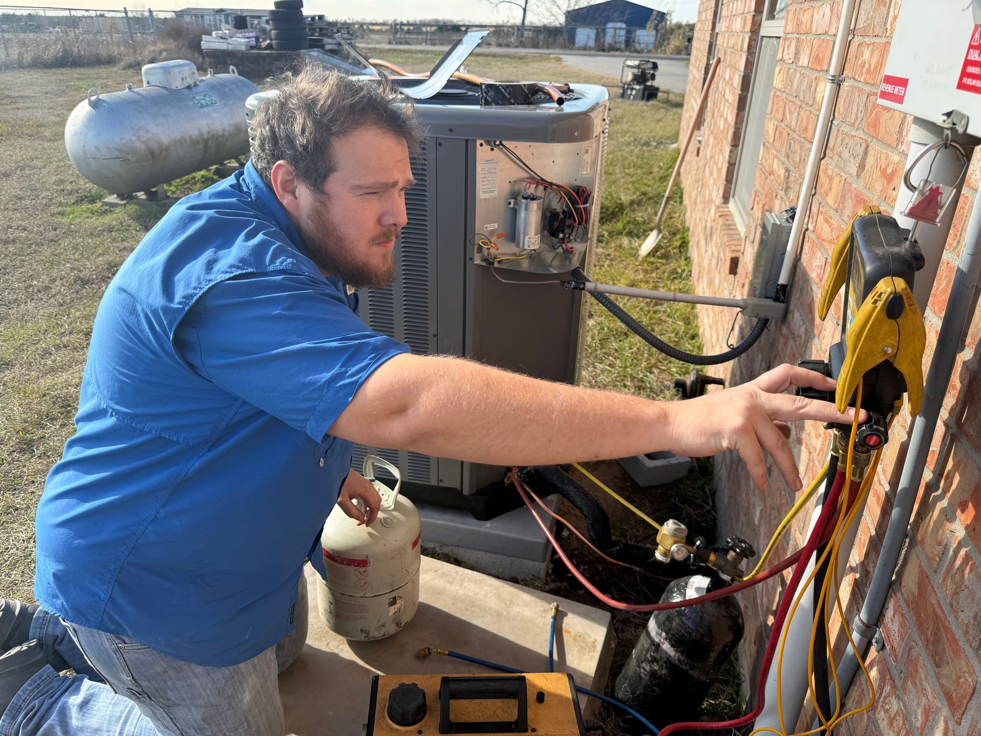 Technician in blue shirt working on outdoor electrical equipment beside wires and tanks.