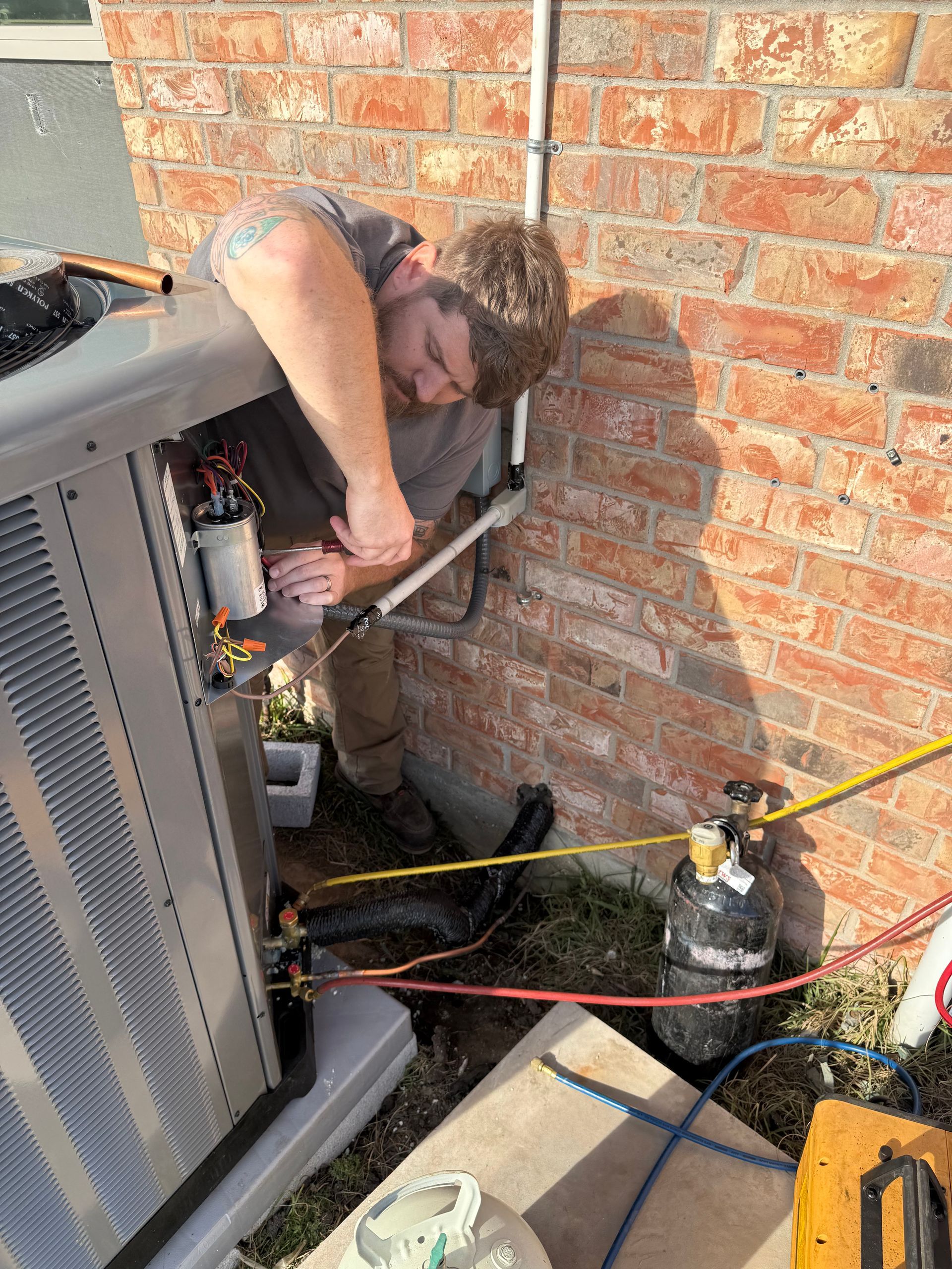 Technician repairing outdoor HVAC unit beside a brick wall, with tools and cables nearby
