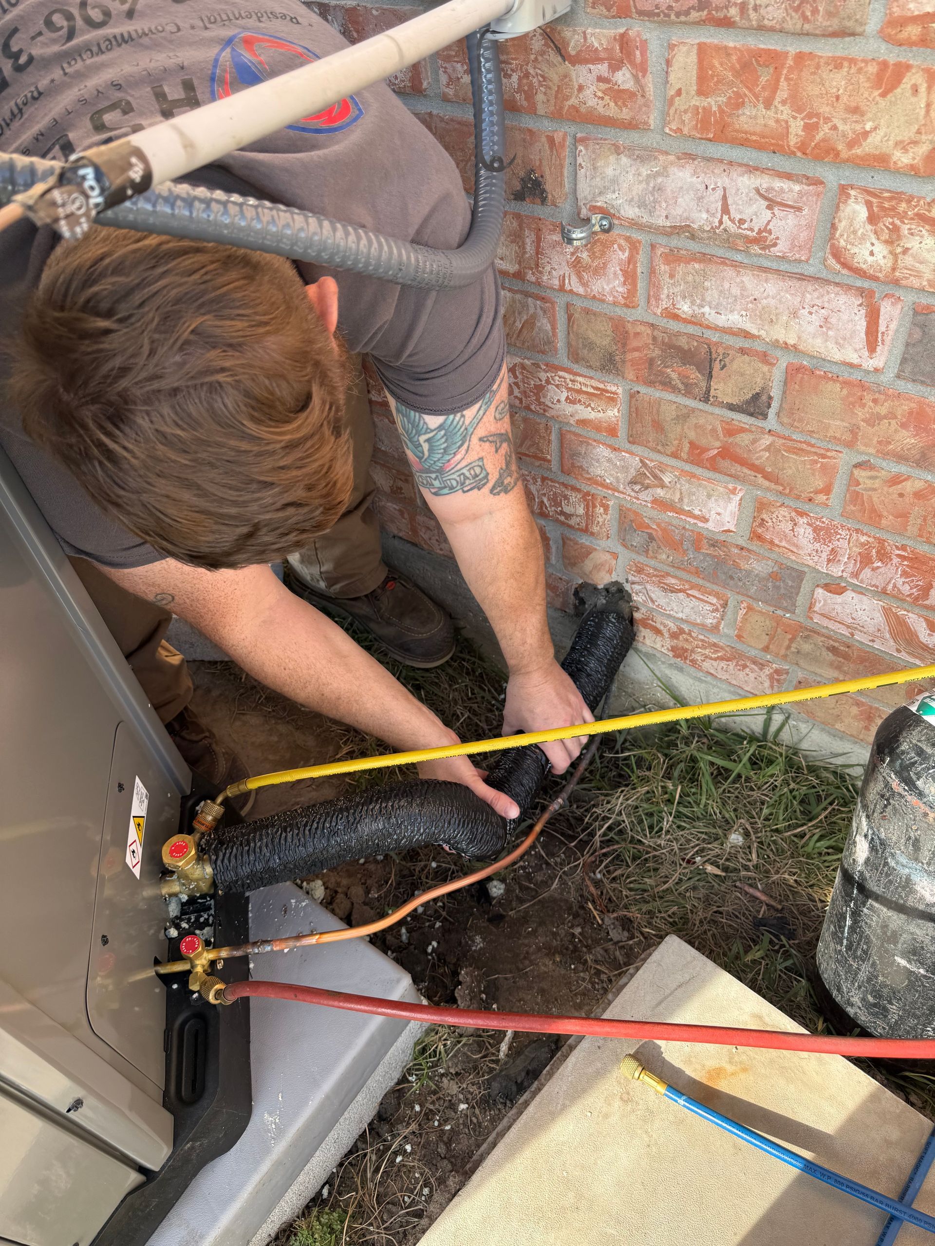 Worker kneeling beside brick wall, inspecting a black trench with measuring tape and cords near HVAC unit