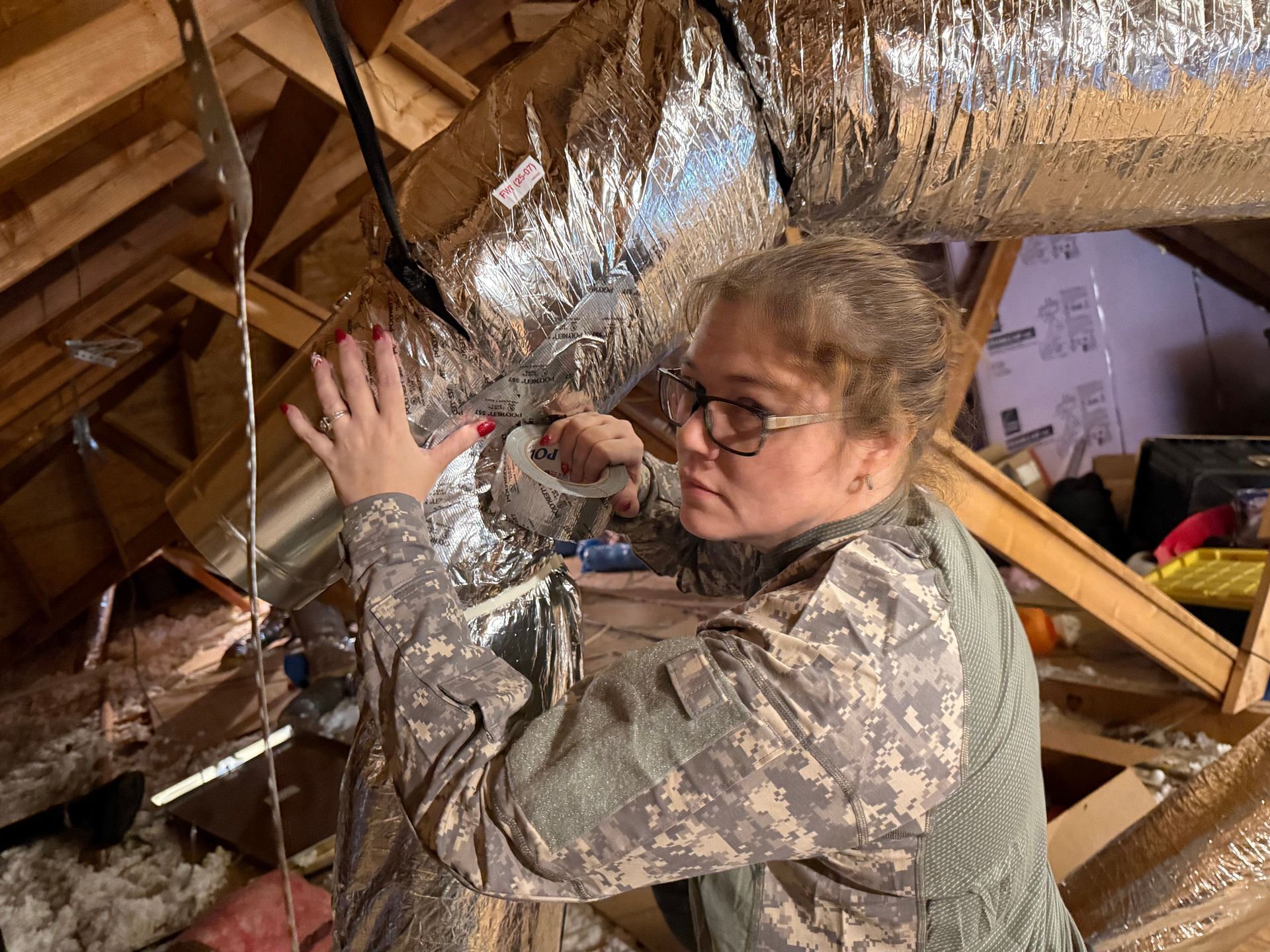 Person in camouflage clothing working in an attic beside shiny duct insulation.