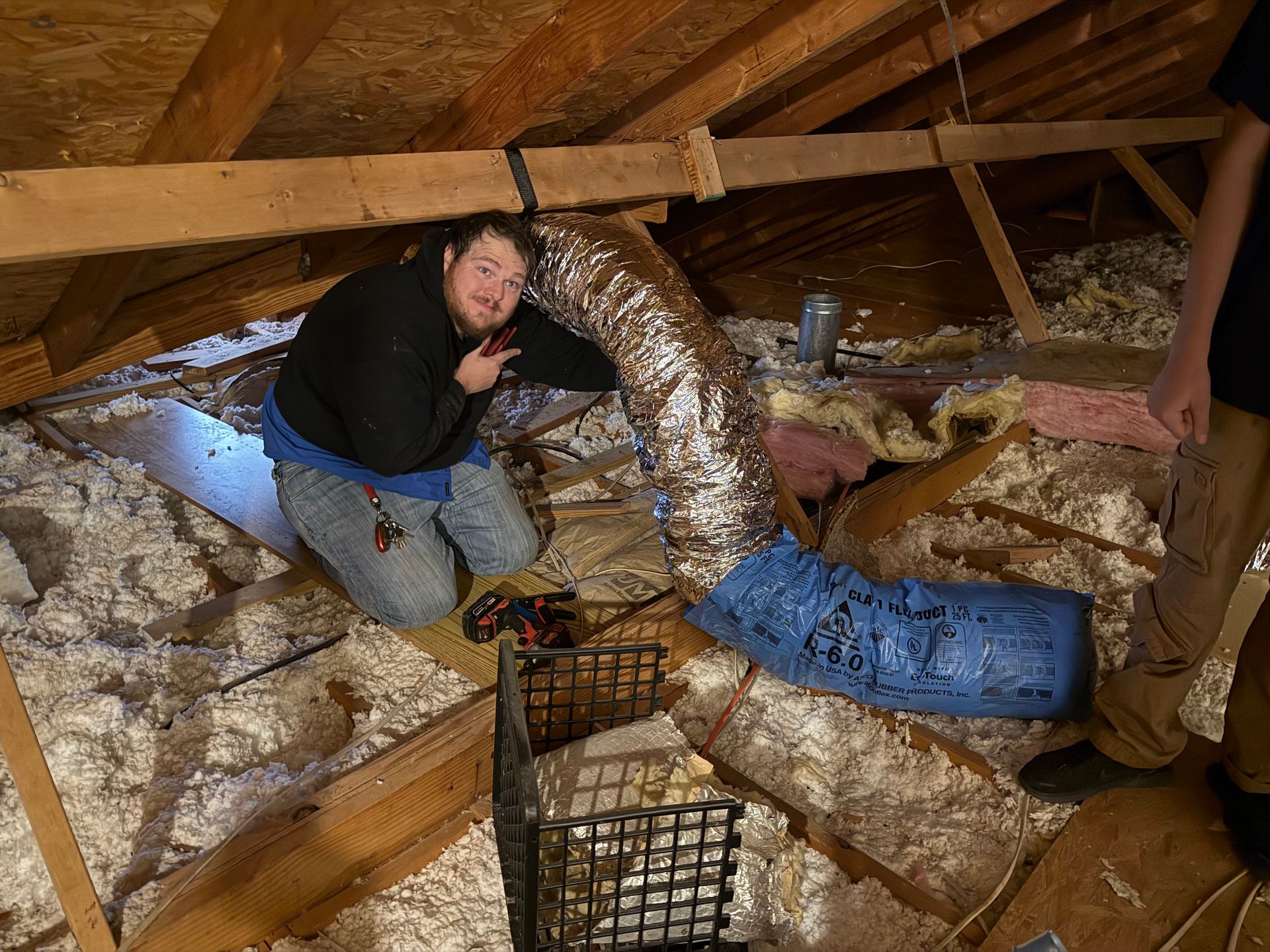 Person kneeling in an attic beside ductwork and insulation, inspecting a floor opening