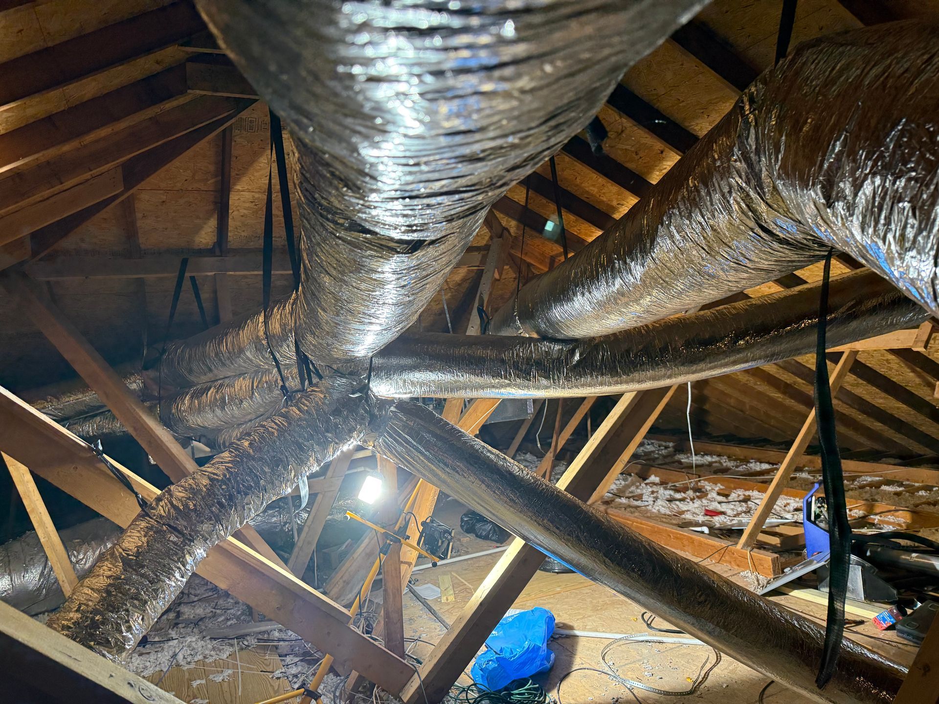 Attic with insulated ductwork, exposed wooden beams, and a bright work light near the center.