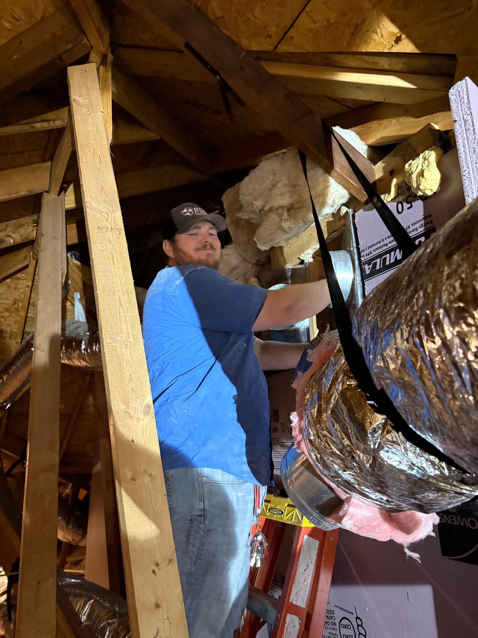 Worker in a wooden attic working on insulated ducting beside a ladder