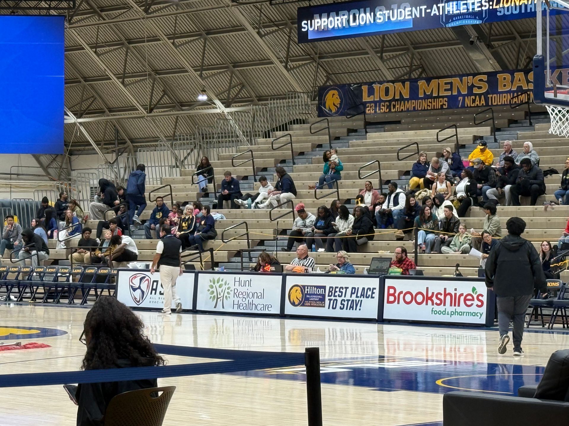 Indoor basketball arena with spectators in the stands and a court game in progress.