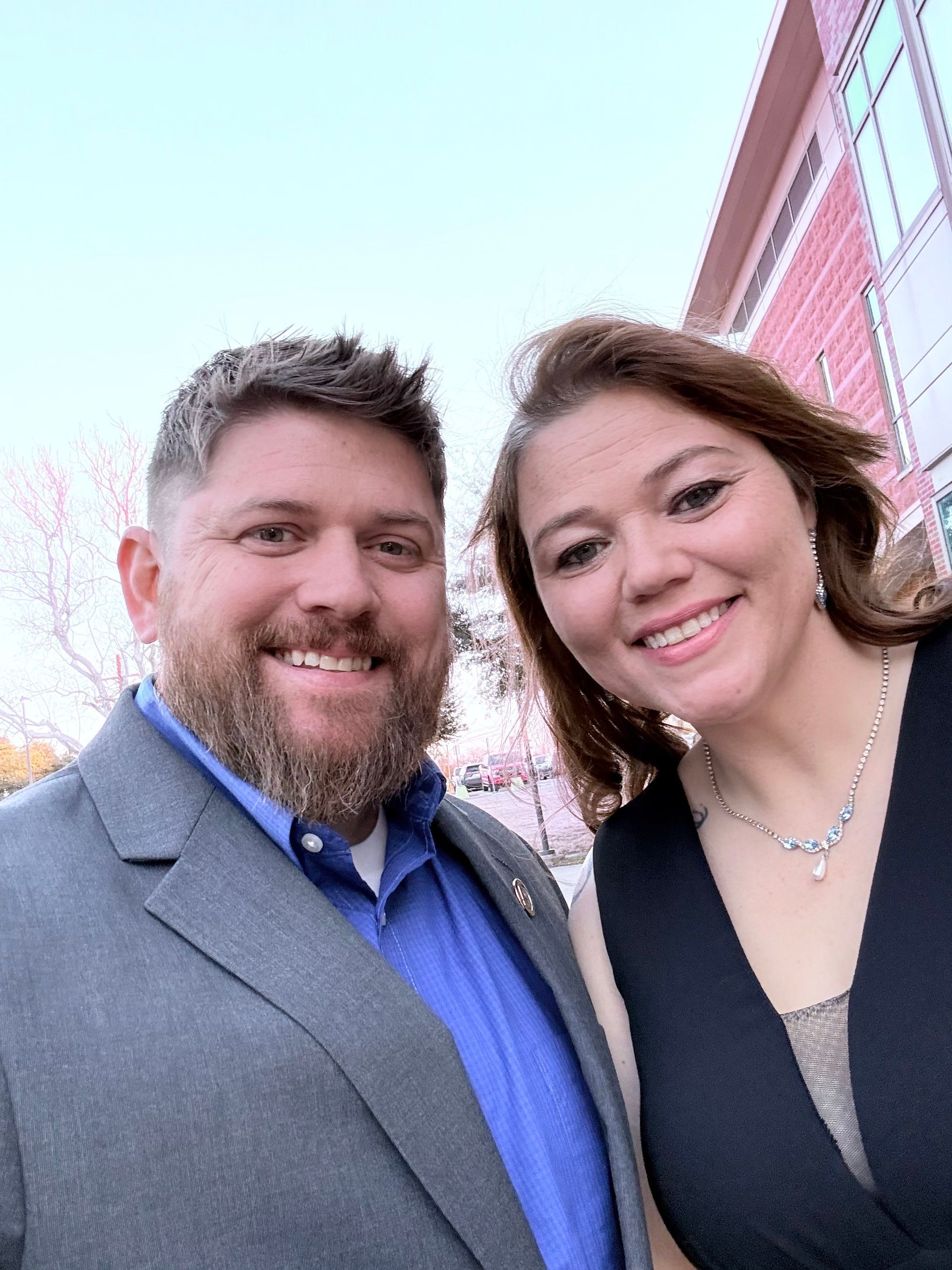 Two people smiling outdoors in front of a red brick building, dressed in formal attire.