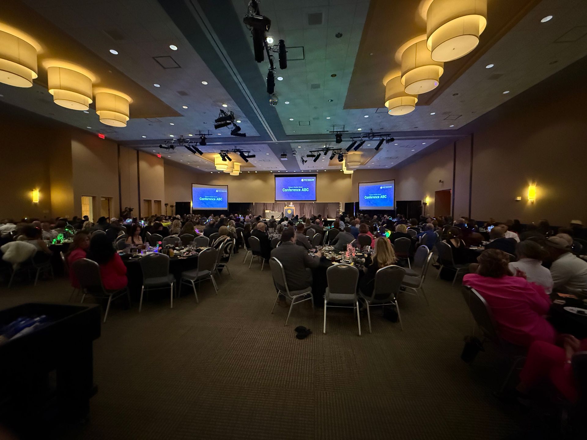 Large conference ballroom with seated audience facing a stage and two blue presentation screens