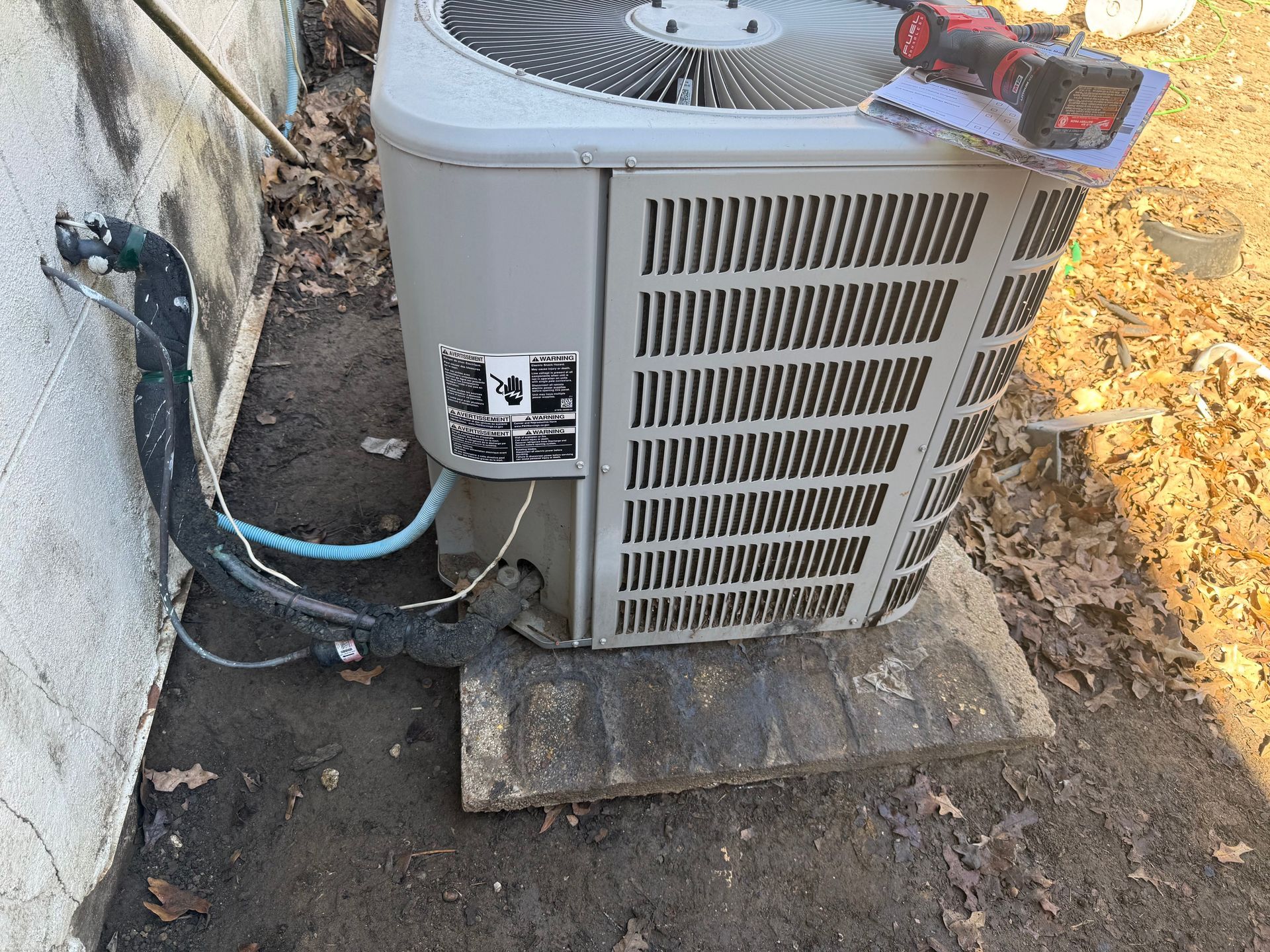 Outdoor air conditioning unit beside a wall, with hoses and tools on top, set on a small concrete pad.
