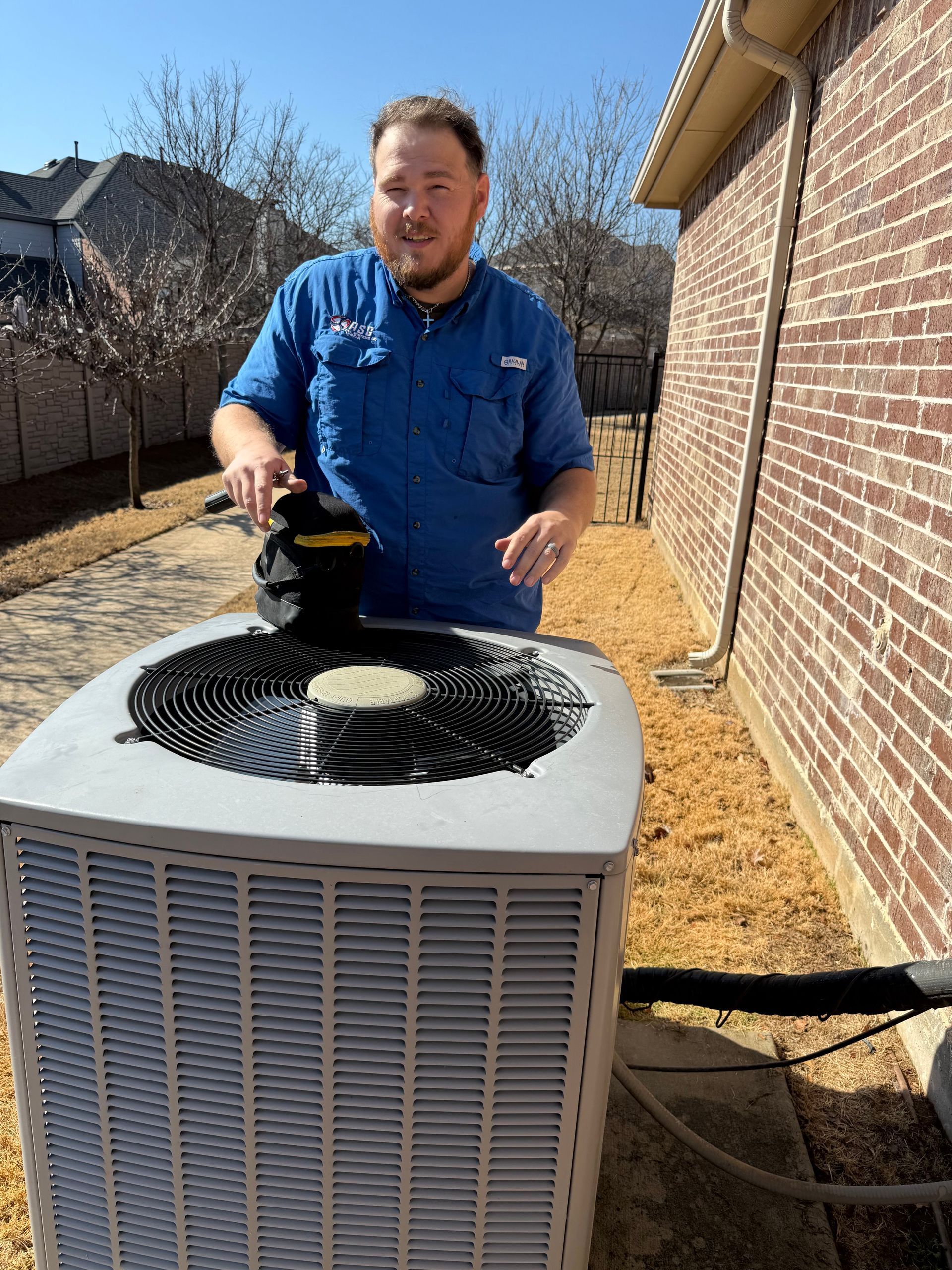 Man using a paintbrush on the top of an outdoor air conditioner unit beside a brick house.