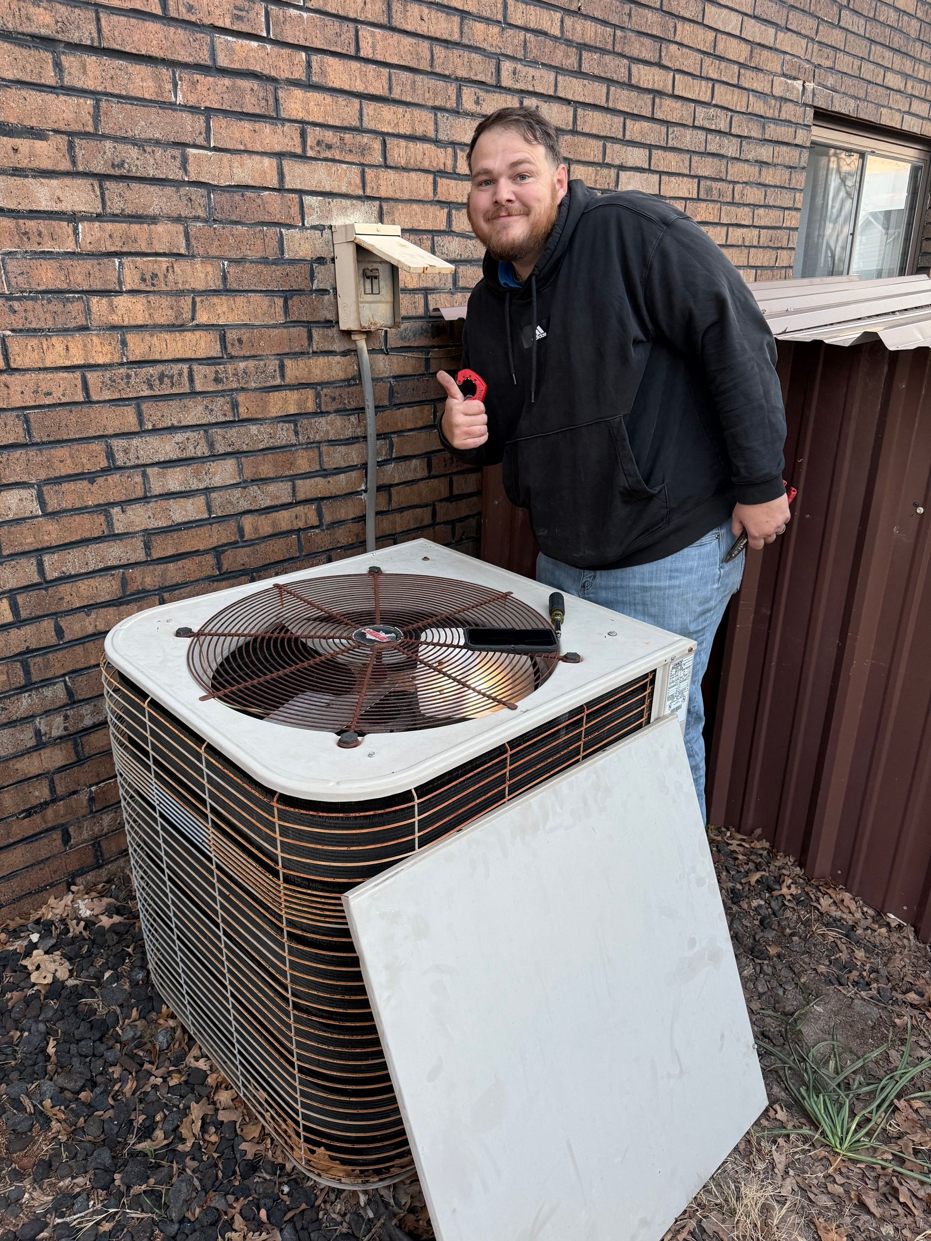 Person crouching beside a rooftop HVAC unit next to a brick wall, holding a tool and smiling.
