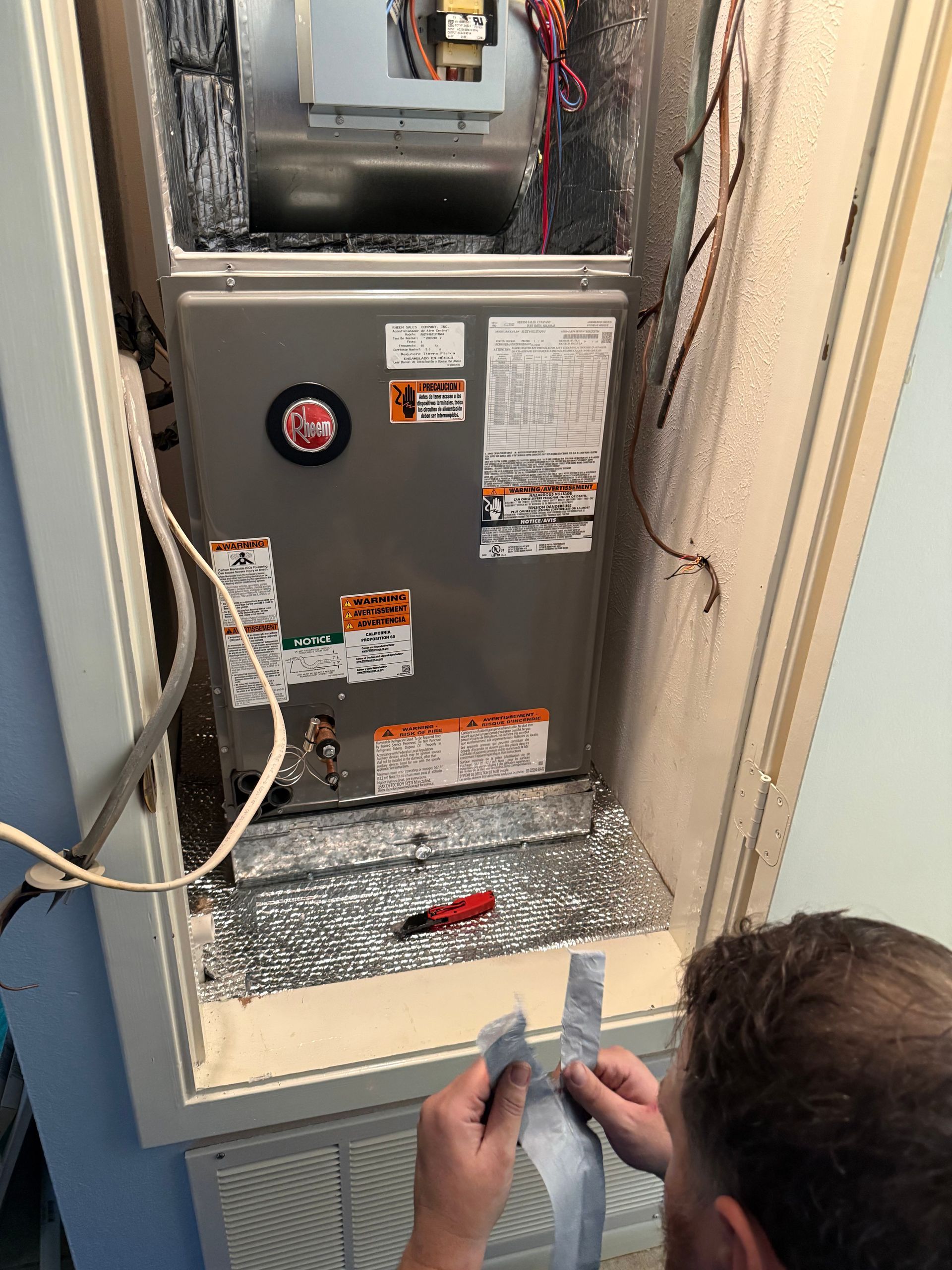 Person holding a tool in front of an open HVAC furnace in a utility closet