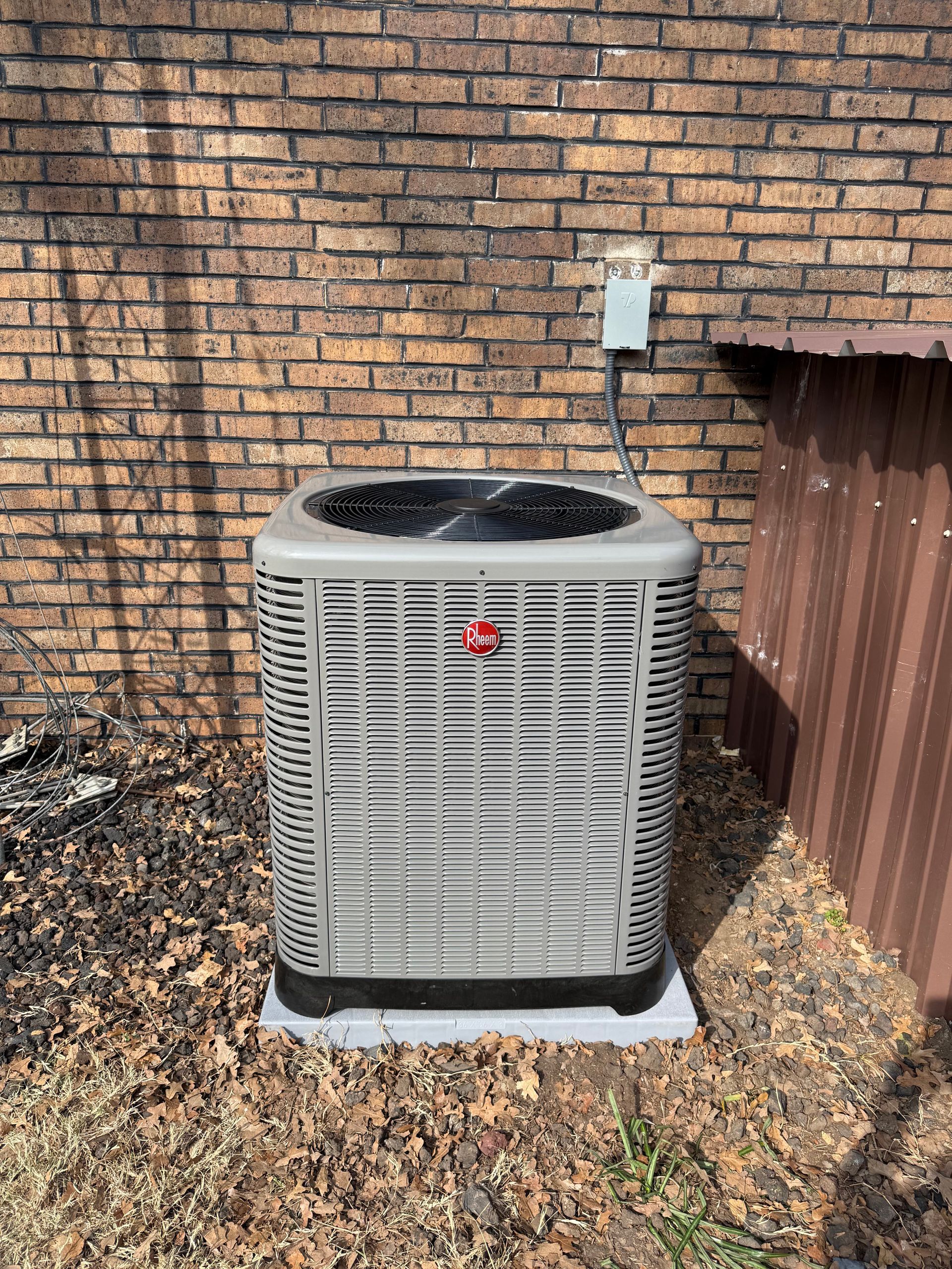 Outdoor HVAC condenser unit beside a brick wall and wooden fence on a leaf-covered ground
