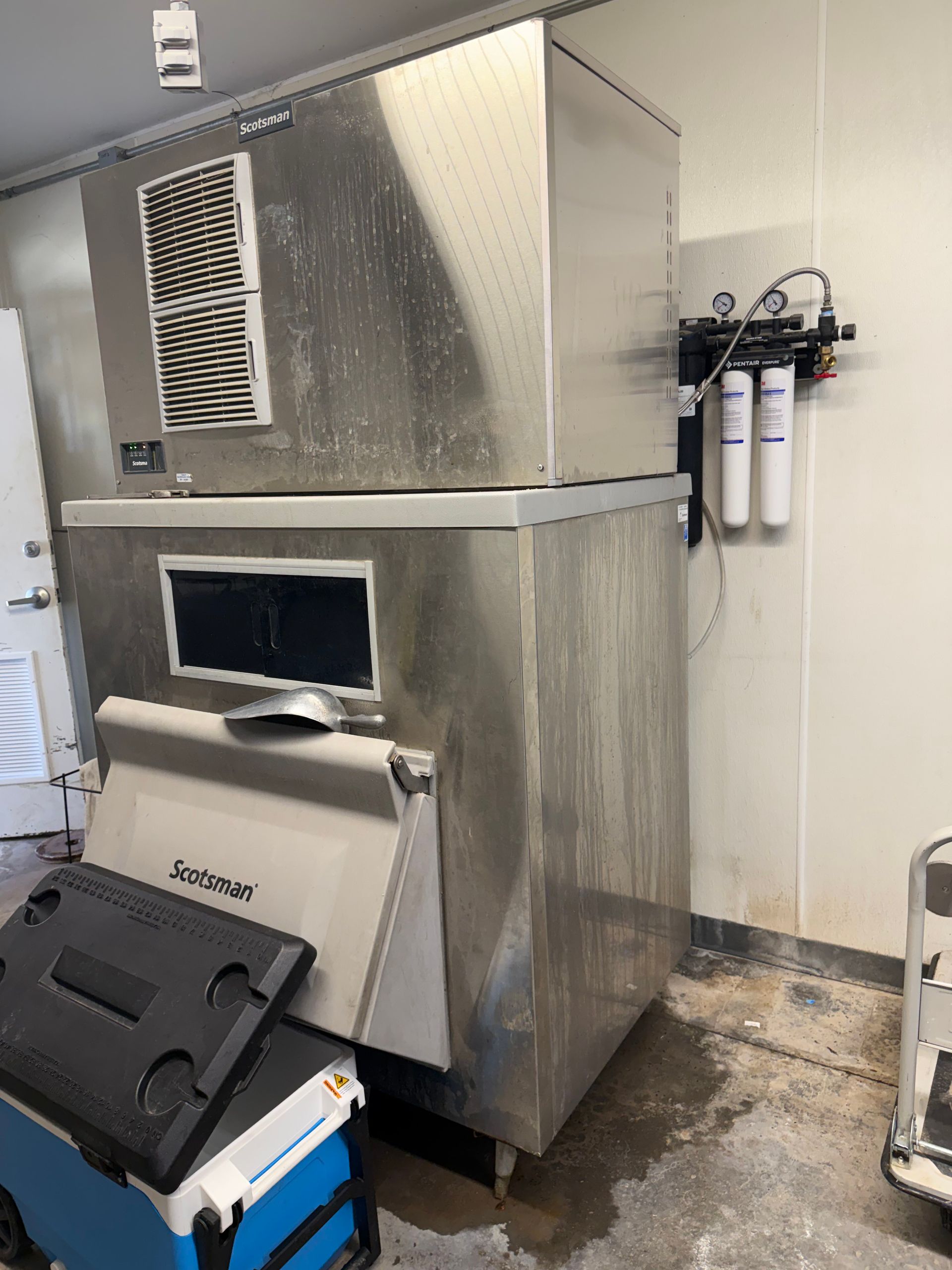 Stainless steel commercial ice machine with storage bin in a utility room, with wall-mounted filters nearby