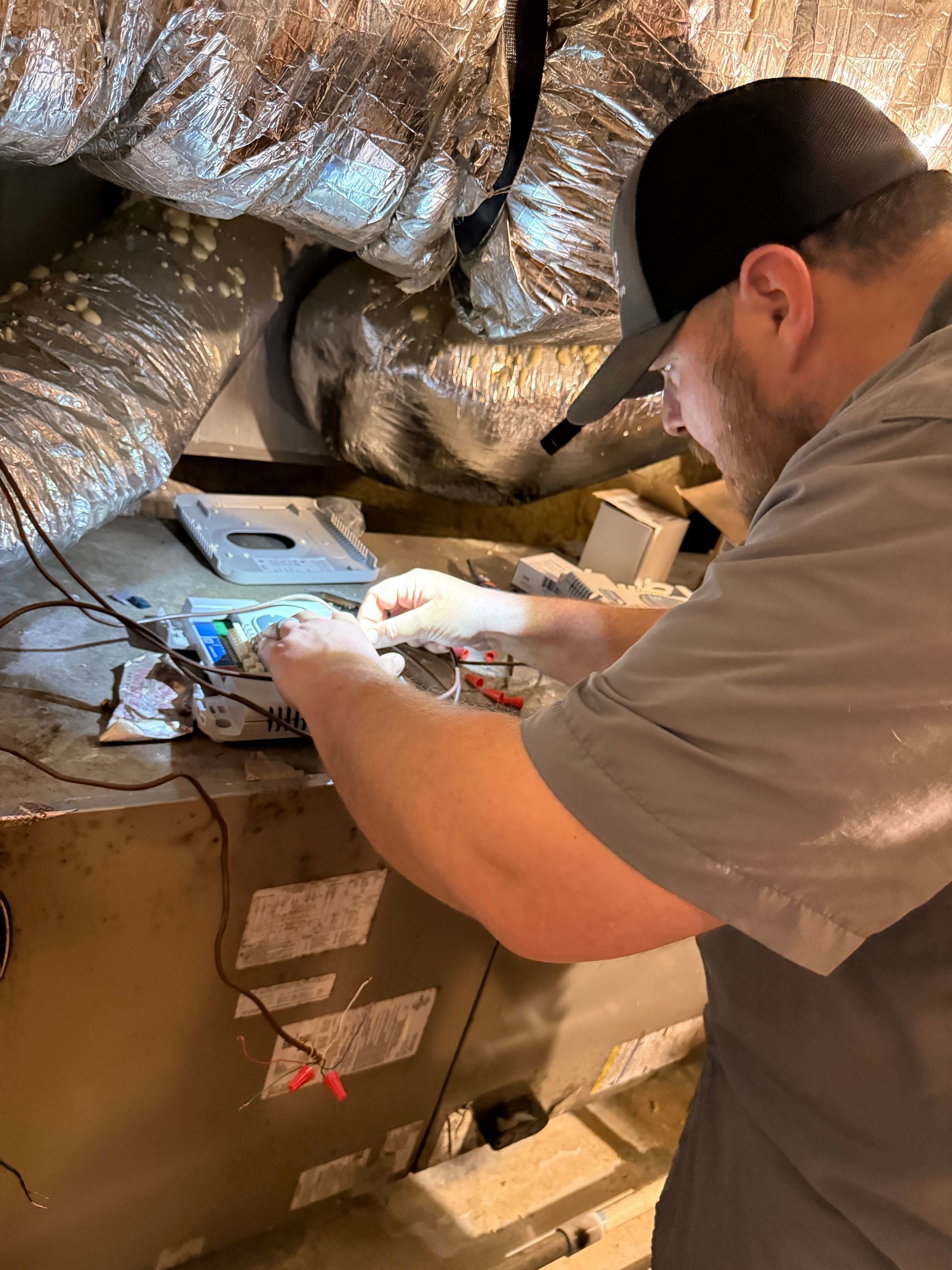 Technician working on exposed wiring inside an attic beside silver ductwork and a control box