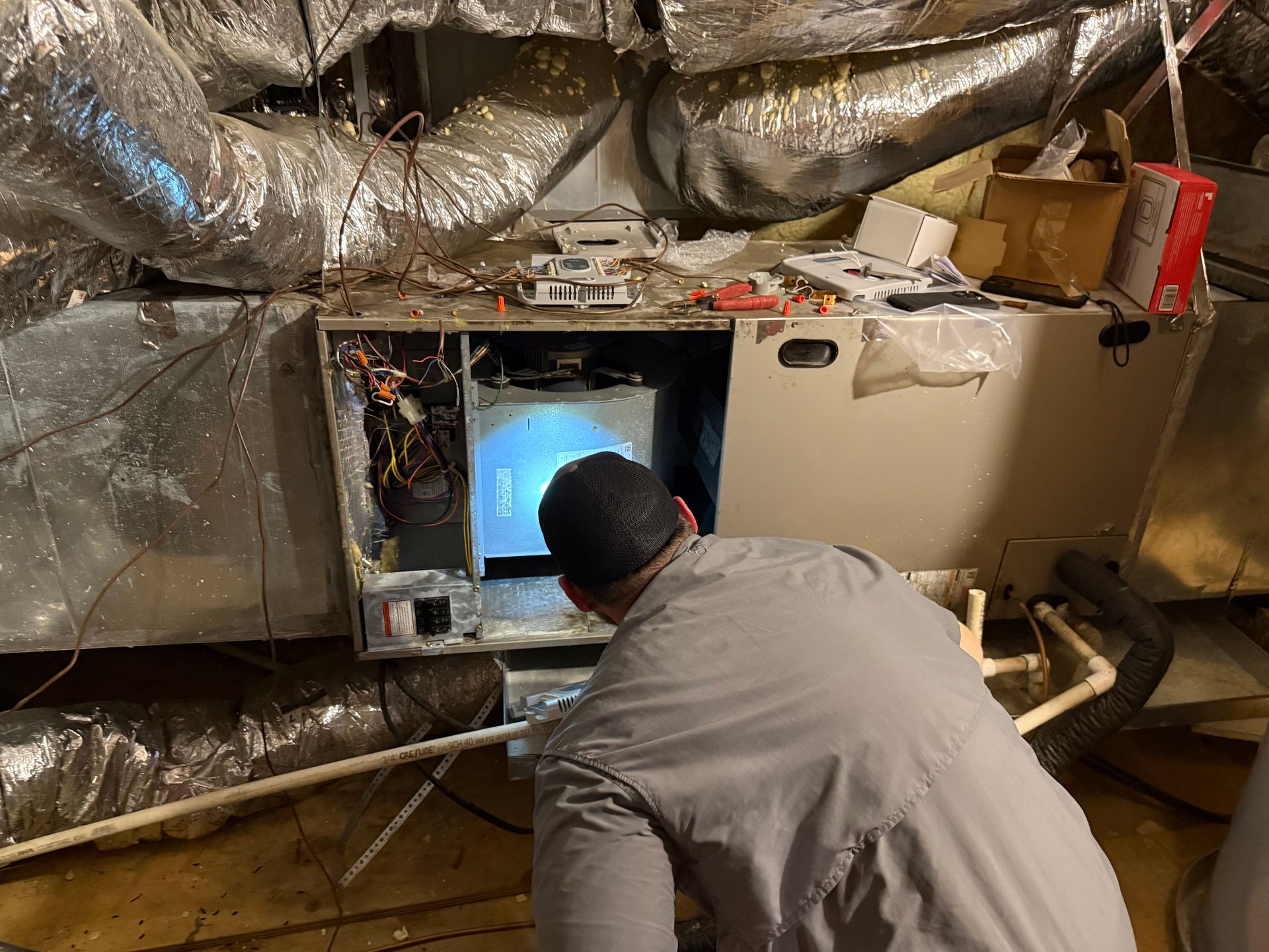 Technician crouches in a basement crawlspace inspecting an HVAC unit amid exposed ductwork and insulation.