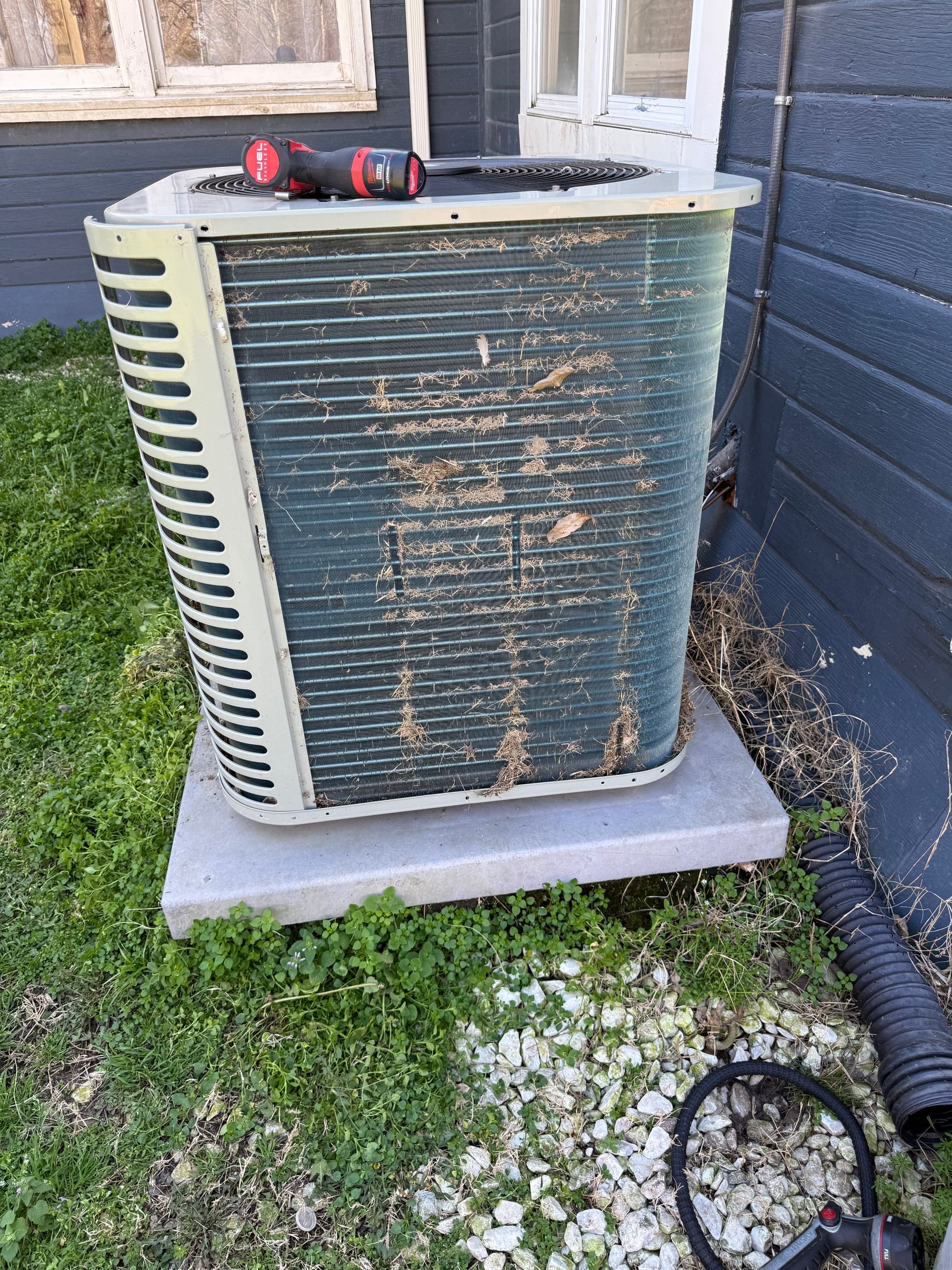 Outdoor air conditioner unit with rusted front grille beside a house wall and window, tools on top